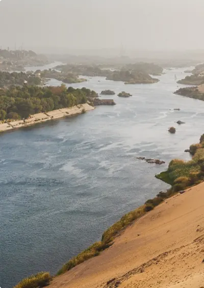 Il Nilo che scorre largo tra rive verdi e dune di sabbia, in una giornata velata